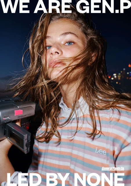 Close-up of a young adult with brown hair holding a camcorder in a night urban setting. Wearing a striped shirt with “Lee” logo.