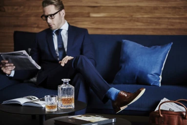 Man in a navy suit wearing glasses reading a newspaper seated on a blue sofa with a whiskey decanter on a table