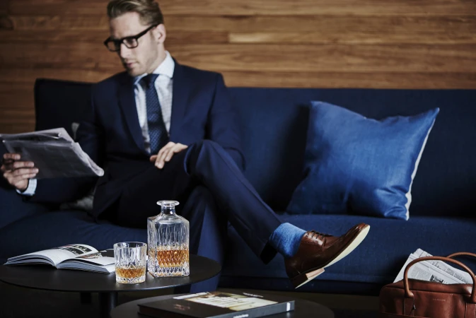 Man in a suit relaxes on a couch, reading a newspaper next to a glass of whiskey and decanter on a coffee table, indoor setting.