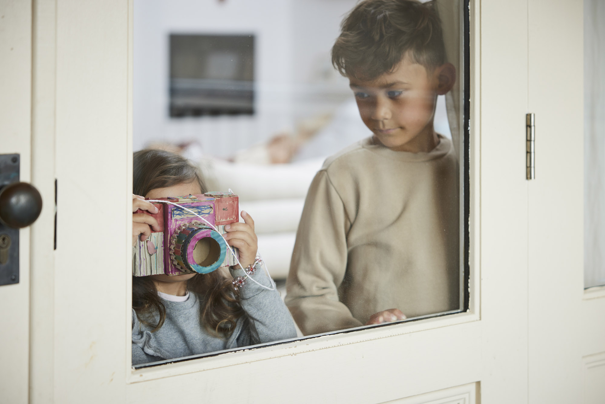 A young girl holds a handmade cardboard camera in front of her face, standing near a boy looking through a white-framed window.