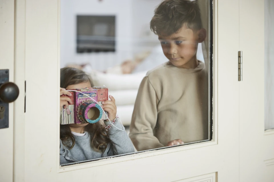 A young girl holds a handmade cardboard camera in front of her face, standing near a boy looking through a white-framed window.