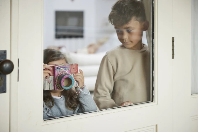 A young girl holds a handmade cardboard camera in front of her face, standing near a boy looking through a white-framed window.