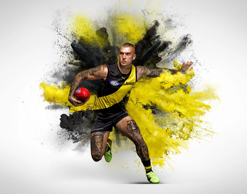 Action shot of a muscular adult male athlete running with a football amidst a burst of black and yellow powder against a white backdrop.