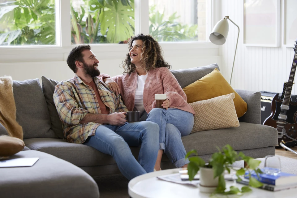 Cheerful couple lounging on gray sofa in brightly lit living room with house plants, guitar, accent pillows, and neutral decor.