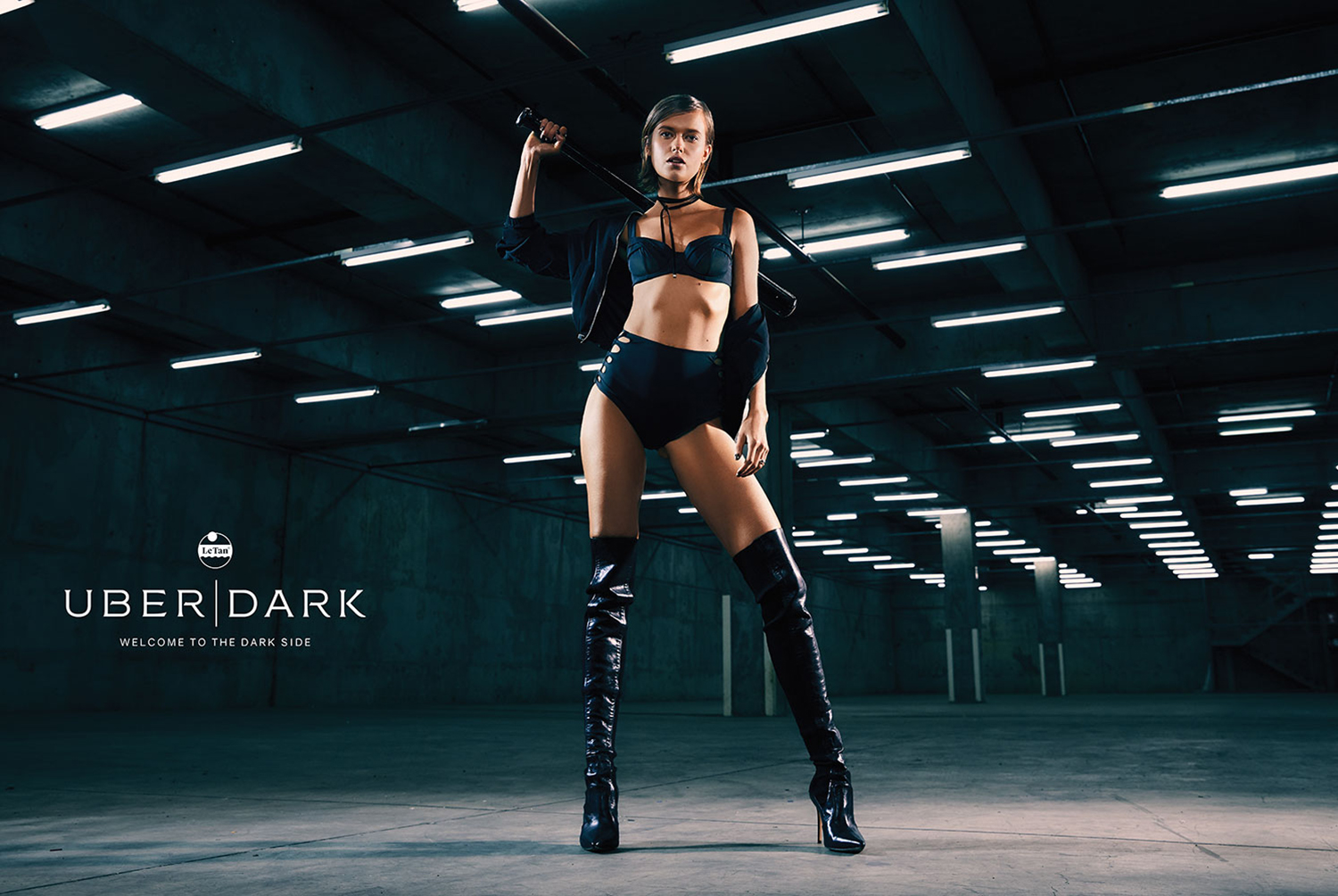 Fashion photo of a slender woman in a black lingerie set and shiny thigh-high boots, holding a baton, in a dark industrial setting.