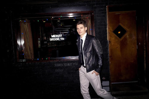 Adult male poses in leather jacket and casual pants in front of a dimly lit building exterior with a window and door.