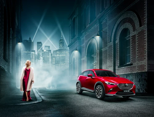 Red Mazda CX-3 parked on city street next to woman in red dress and white coat, cityscape in background, night scene, streetlights.