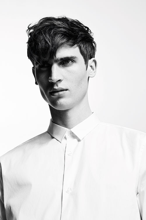 Black and white portrait of an adult male with dark, wavy hair, wearing a light-colored button-down shirt against a bright background.