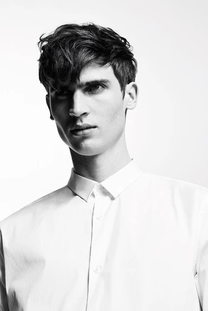 Black and white portrait of an adult male with dark, wavy hair, wearing a light-colored button-down shirt against a bright background.