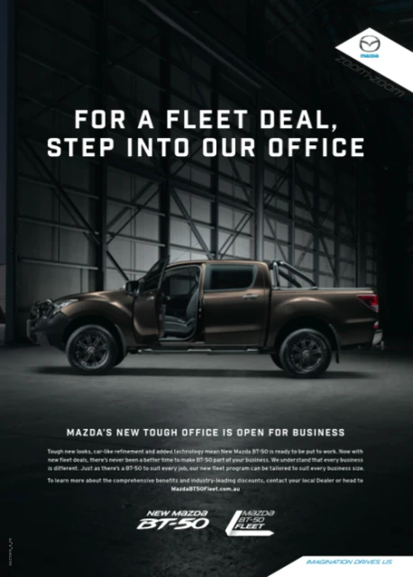 Brown Mazda BT-50 pickup truck parked inside a warehouse-style building, spotlighted with an open driver door.