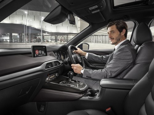 Adult male driving black Mazda CX-9 SUV with leather interior. He wears a gray suit, and city buildings are visible in the background.