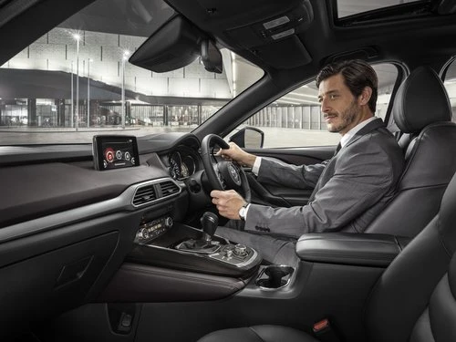 Adult male driving black Mazda CX-9 SUV with leather interior. He wears a gray suit, and city buildings are visible in the background.