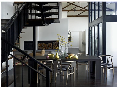 Interior view of a modern dining room with a dark wood table, wishbone chairs, a fireplace, and black metal stairs in a contemporary home.
