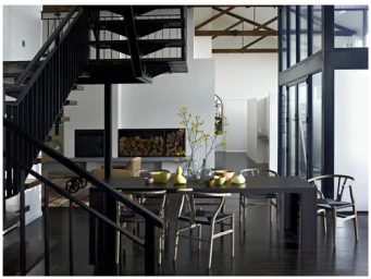 Modern open-plan interior featuring a dark dining table, wooden chairs, glass vases with yellow stems, and a black staircase.