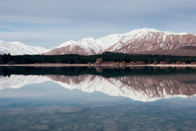 Snow-capped mountain range reflected in a calm mirror-like lake with a dark forest line along the shore under an overcast sky.