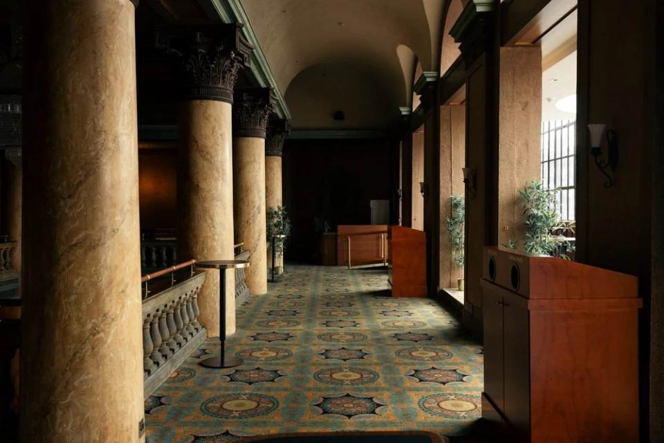 Ornate hallway with columns and geometric patterned carpet leading to reception desks and natural light in a grand indoor space.