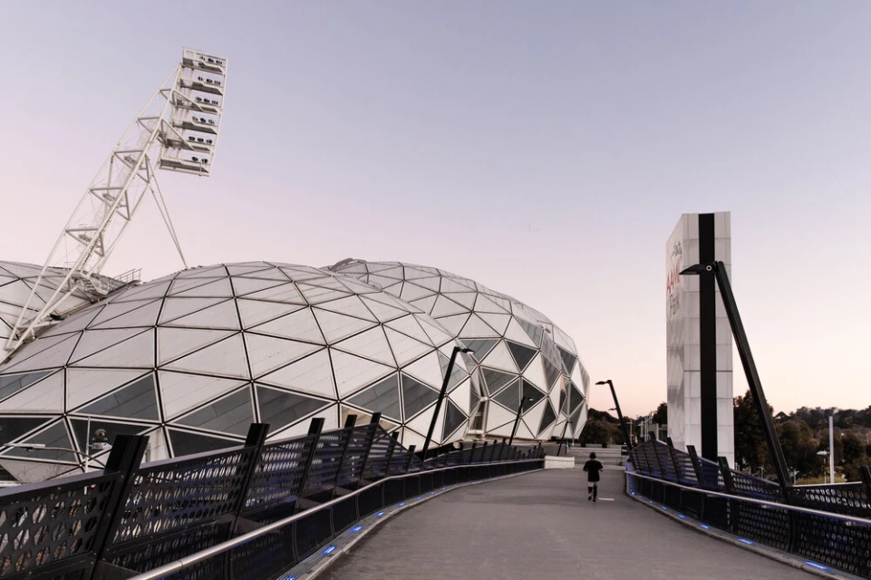 Exterior view of Marvel Stadium in Melbourne, Australia at dusk with its dome-shaped roof, lights, and walkway; a person runs along the walkway.