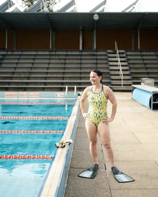 Woman in swimsuit and swim fins stands poolside by a pool at an outdoor swimming venue.