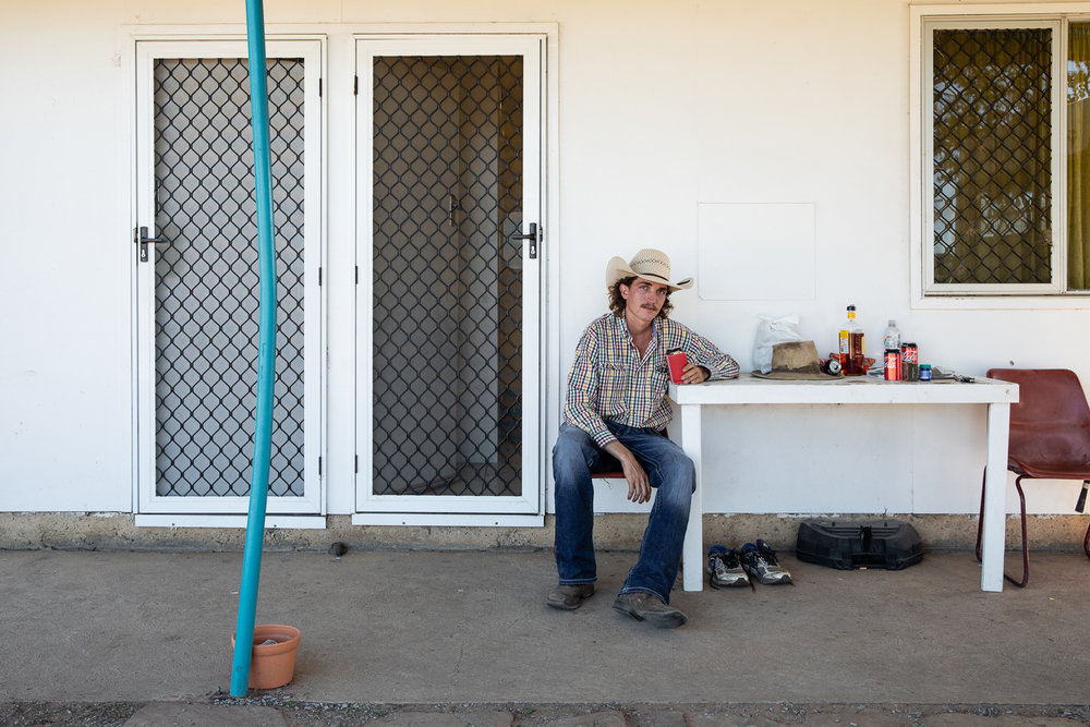 Adult man in a cowboy hat sits on a chair and drinks at a white table outside near doors on a white building in a daylight scene.
