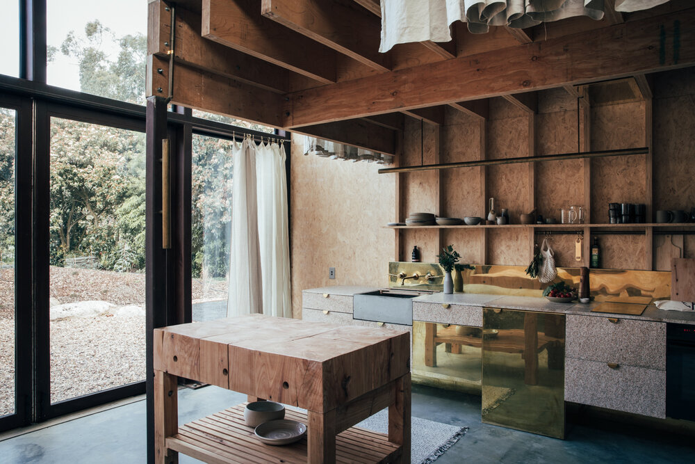 Modern plywood kitchen interior featuring a butcher block island, brass details, and natural light from a large window overlooking greenery.