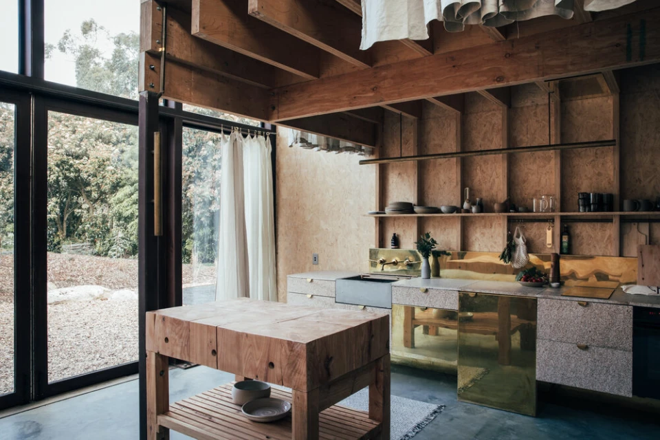 Modern plywood kitchen interior featuring a butcher block island, brass details, and natural light from a large window overlooking greenery.