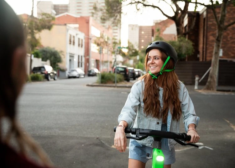 Young woman with long auburn hair smiles while riding an electric scooter on a city street wearing a helmet and denim jacket.
