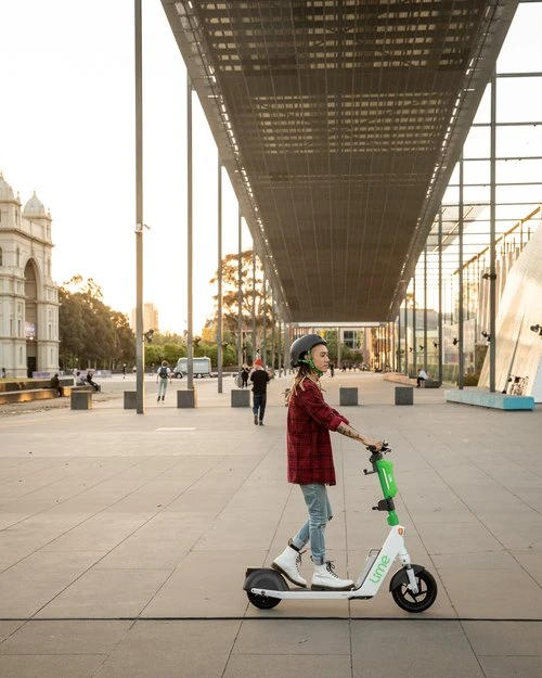 Person rides Lime electric scooter on city square near building with archways on a sunny day in an urban environment with few people.