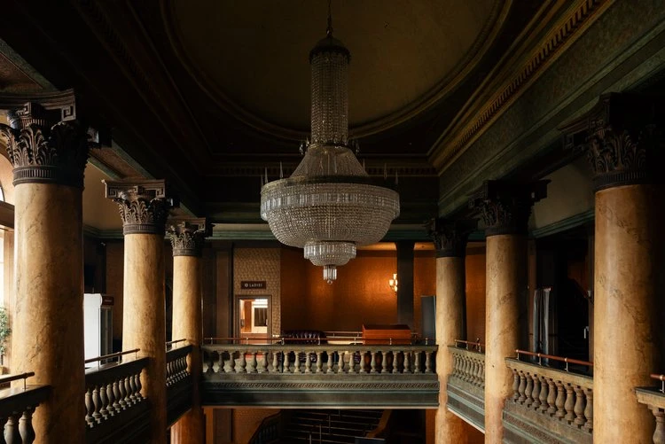 Ornate theatre lobby interior with columns, crystal chandelier, and balcony under dim lighting, creating a dramatic architectural space.