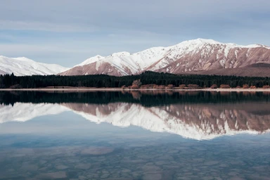 Snow-capped mountain range reflected in a calm mirror-like lake with a dark forest line along the shore under an overcast sky.