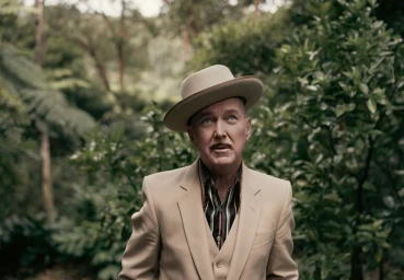 Man wearing a cream suit and fedora hat standing outdoors among green trees and lush foliage