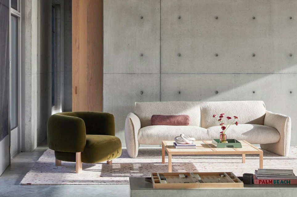 Interior living space features a beige sofa with a velvet lumbar pillow, an olive green lounge chair, a wood table, and a concrete wall.