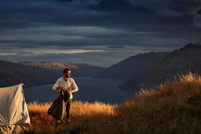 Bearded man in white henley standing in tall grass beside a tent, overlooking a lake and mountain range under a dramatic dark sky.
