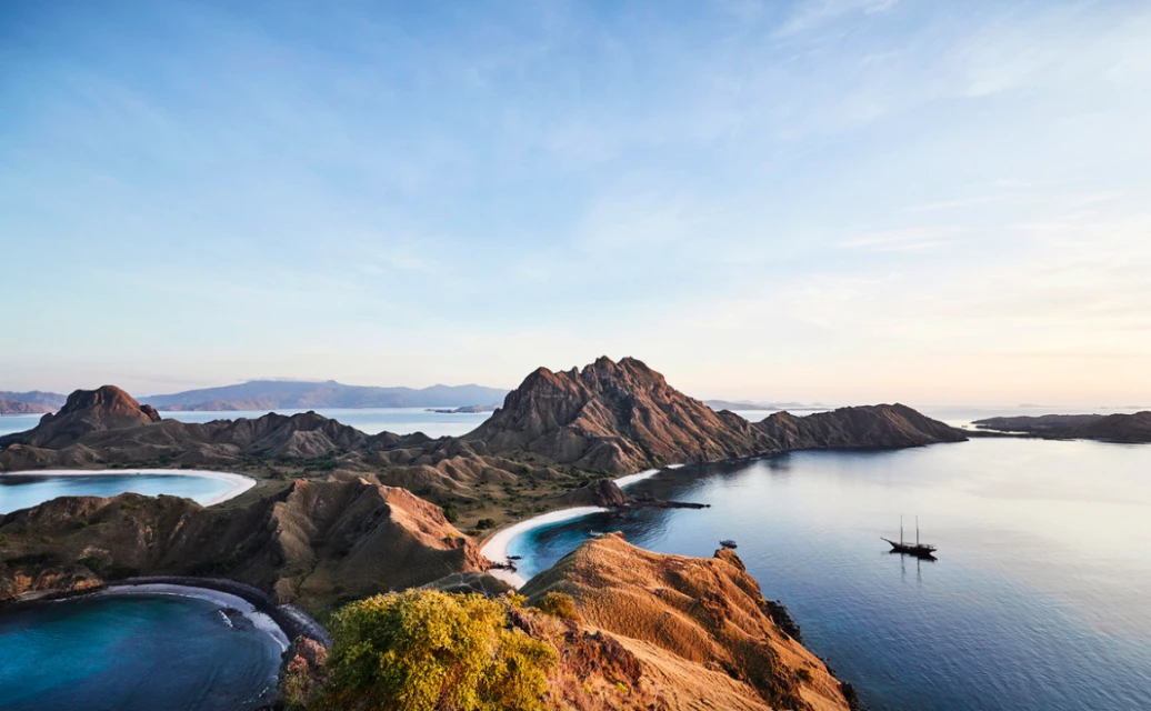 Coastal mountains meet turquoise sea in a scenic panorama; island landscape with a sailboat gliding across the water under a blue sky.