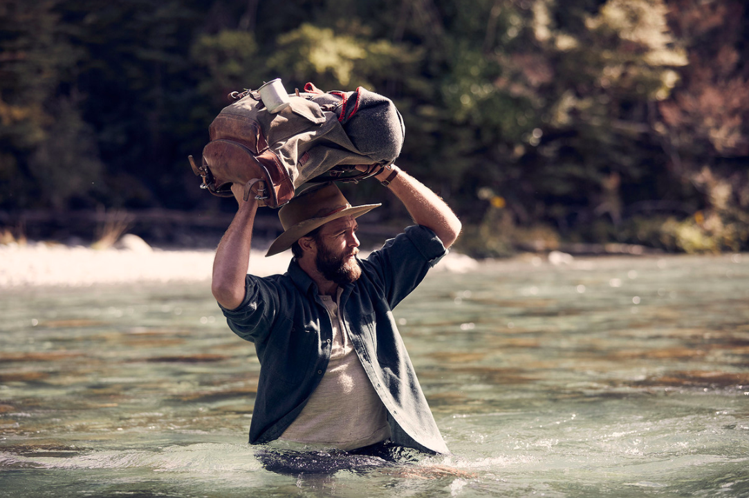 Adult male wading in a river, carrying a large backpack overhead, wearing a hat and blue shirt with a forest background.