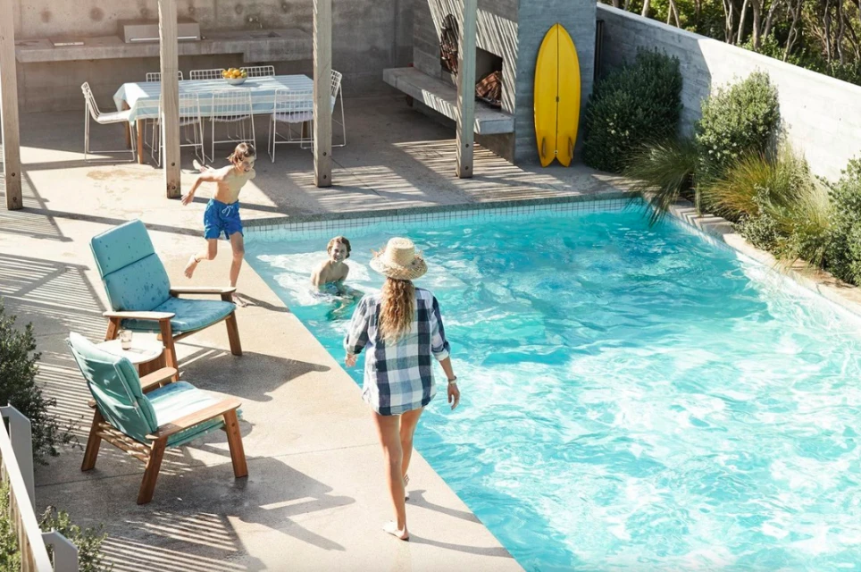 Outdoor scene features a woman walking by a pool with children playing, chairs, table and surfboards in the background, and greenery.