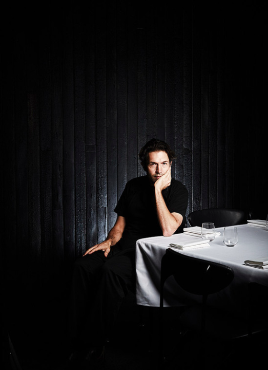 Man in a black shirt sitting at a table set with plates and glasses in a darkly lit restaurant with a black wood-paneled wall.