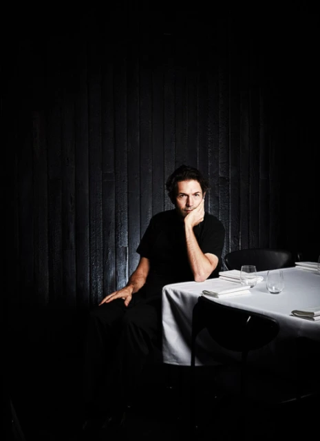 Man in a black shirt sitting at a table set with plates and glasses in a darkly lit restaurant with a black wood-paneled wall.