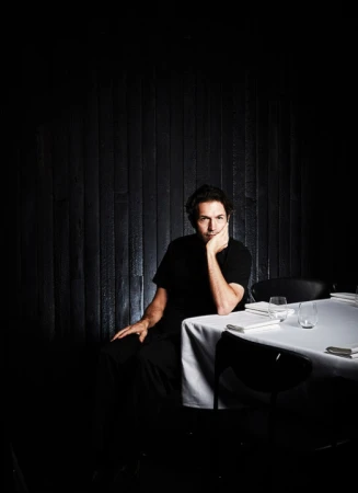 Man in a black shirt sitting at a table set with plates and glasses in a darkly lit restaurant with a black wood-paneled wall.