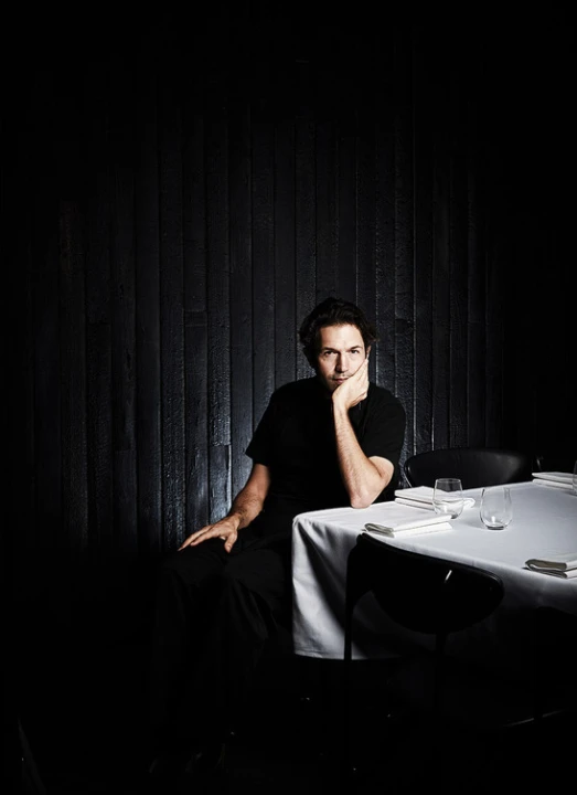 A man in a black t-shirt seated at a white-clothed table against a dark slatted wall, photographed by James Geer.