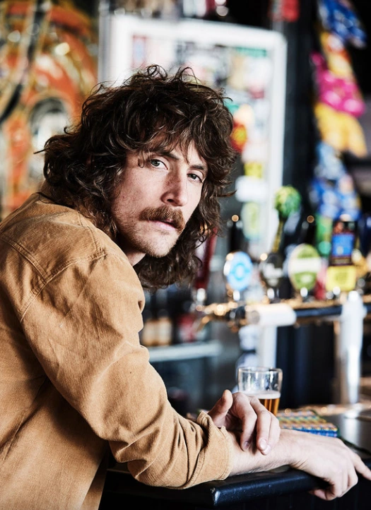Man with curly brown hair and a mustache wearing a tan jacket sitting at a bar counter with beer taps in the background.