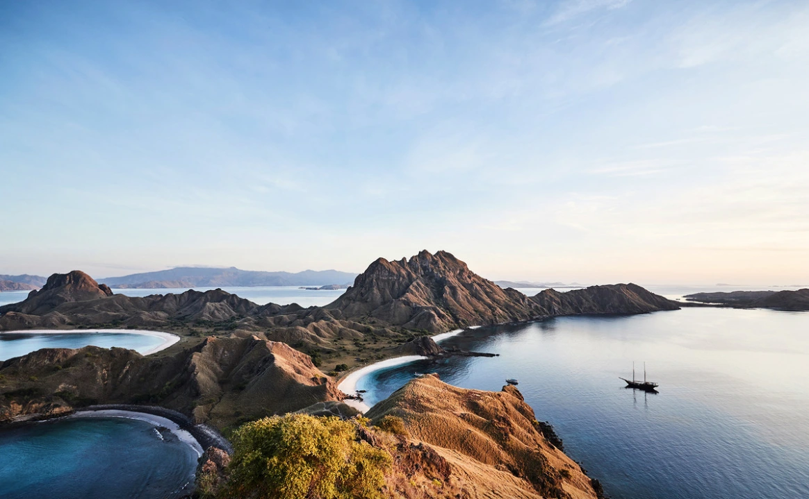 High-angle aerial view of rugged coastal mountains and sandy beaches surrounding calm blue ocean water with a sailing ship