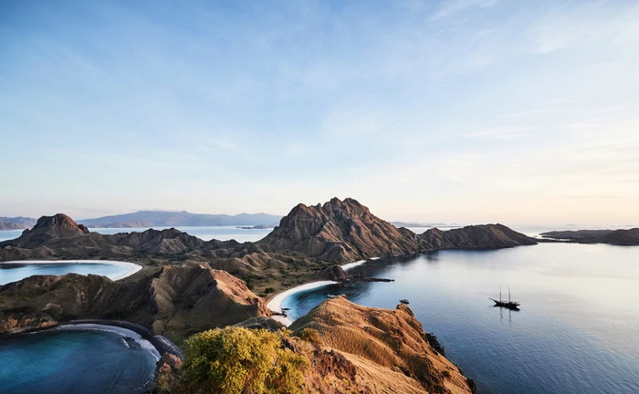 High-angle aerial view of rugged coastal mountains and sandy beaches surrounding calm blue ocean water with a sailing ship