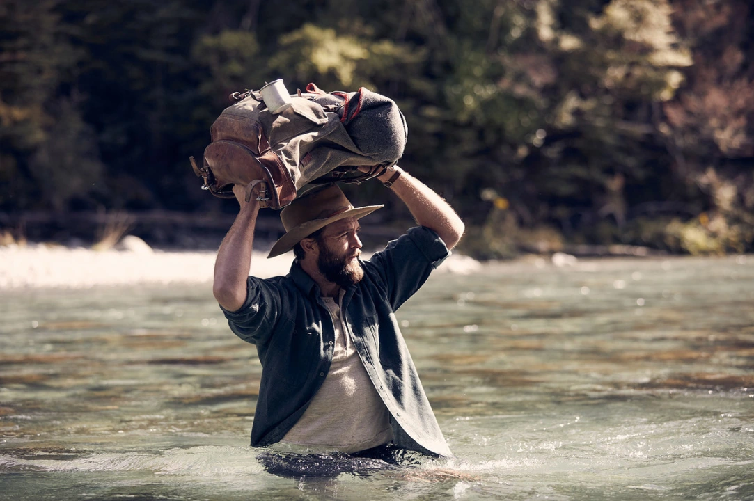 Bearded man wading through a river while holding a large canvas backpack above his head