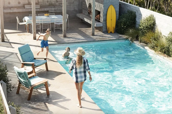 Woman walking toward a sunny backyard swimming pool with two children playing while a yellow surfboard leans against a concrete wall.