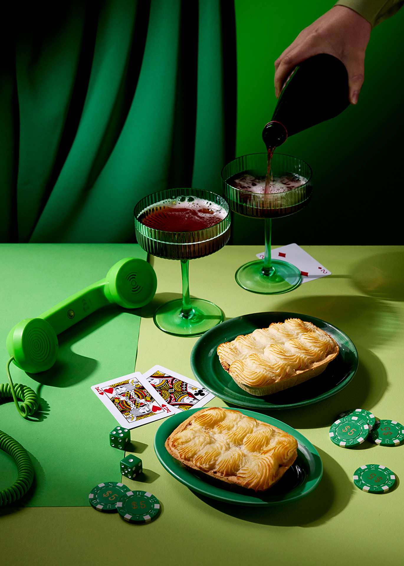 Retro-themed table setting with food, beverages, and gaming elements in shades of green, featuring pies, cocktails, phone and playing cards.