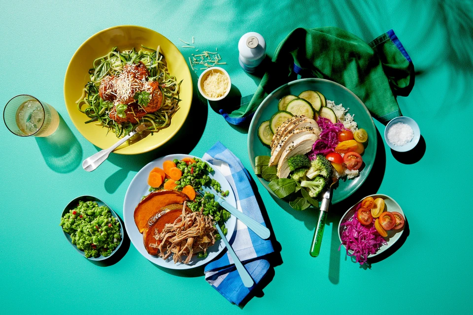 Overhead shot of curated plates and bowls of healthy meal prep, including pasta, protein, grains, and vegetables on a teal surface.