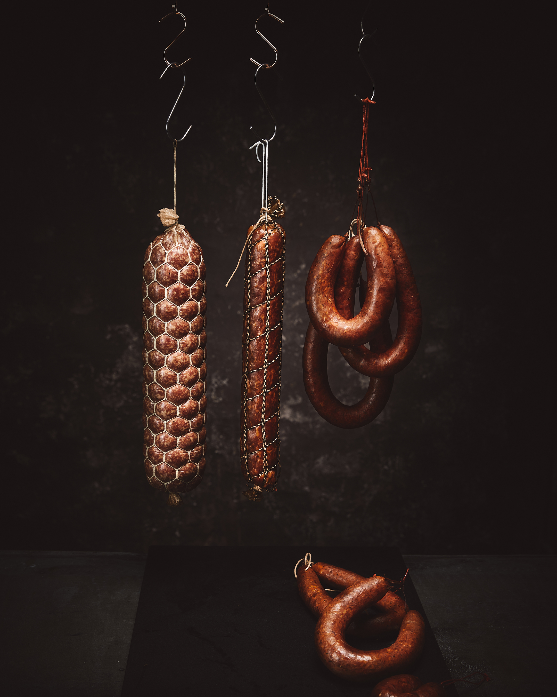 Three types of sausages hang from metal hooks against a dark background, one laying at the bottom on a black surface.
