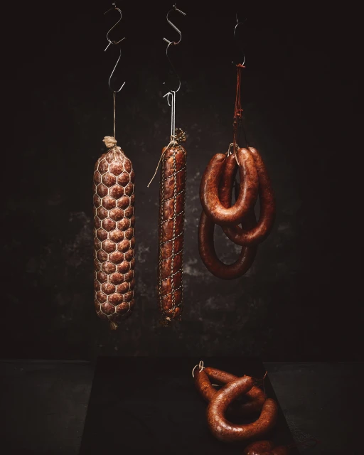 Three types of sausages hang from metal hooks against a dark background, one laying at the bottom on a black surface.