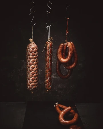 Three types of sausages hang from metal hooks against a dark background, one laying at the bottom on a black surface.