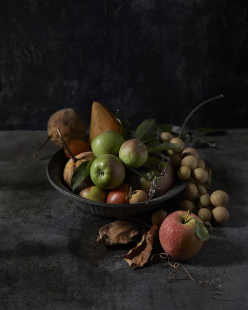 Still life featuring a bowl of apples, pears, longan fruit, and autumn leaves against a dark background with studio lighting.